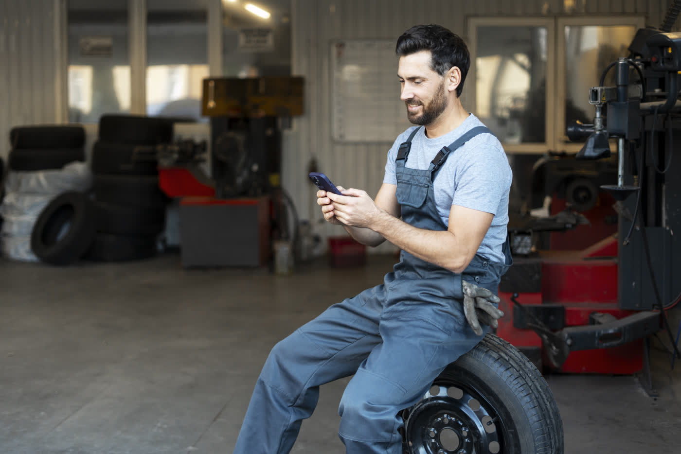 A mechanic sitting on a tire in an auto shop, phone in hand